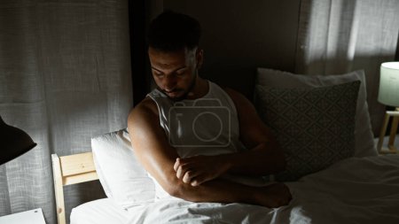 Téléchargez les photos : Jeune homme arabe assis sur le lit bras à gratter pour les démangeaisons à la chambre - en image libre de droit