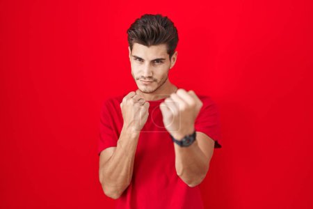 Photo for Young hispanic man standing over red background ready to fight with fist defense gesture, angry and upset face, afraid of problem - Royalty Free Image