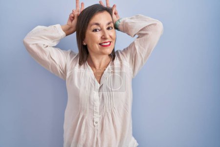 Foto de Mujer hispana de mediana edad de pie sobre fondo azul posando divertida y loca con los dedos en la cabeza como orejas de conejo, sonriendo alegre - Imagen libre de derechos