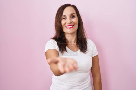 Téléchargez les photos : Moyen âge brunette femme debout sur fond rose souriant joyeuse offrant main de paume donnant assistance et acceptation. - en image libre de droit