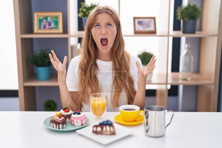 Photo for Young caucasian woman eating pastries t for breakfast crazy and mad shouting and yelling with aggressive expression and arms raised. frustration concept. - Royalty Free Image