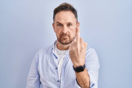 Foto de Middle age caucasian man standing over blue background showing middle finger, impolite and rude fuck off expression - Imagen libre de derechos