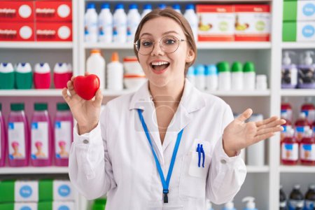 Photo for Young caucasian woman working at pharmacy drugstore holding red heart celebrating achievement with happy smile and winner expression with raised hand - Royalty Free Image