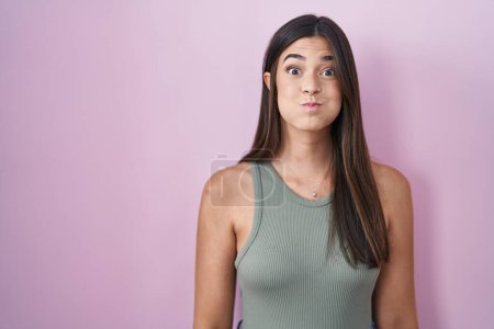 Foto de Mujer hispana de pie sobre fondo rosa hinchando mejillas con cara divertida. boca hinchada de aire, expresión loca. - Imagen libre de derechos