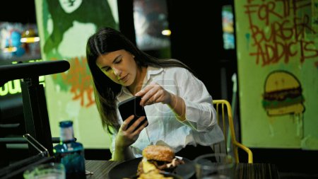 Photo for Young beautiful hispanic woman taking picture of delicious hamburger with smartphone at the restaurant - Royalty Free Image