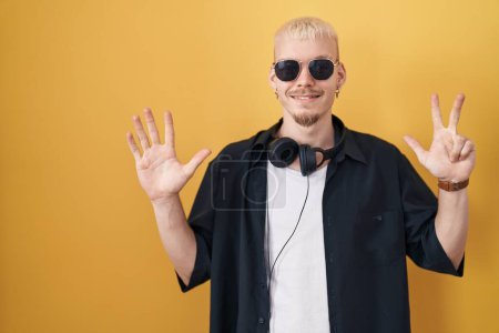 Téléchargez les photos : Jeune homme caucasien portant des lunettes de soleil debout sur fond jaune montrant et pointant vers le haut avec les doigts numéro huit tout en souriant confiant et heureux. - en image libre de droit