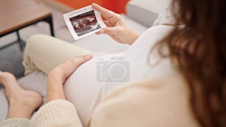 Photo for Young pregnant woman sitting on sofa looking baby ultrasound at home - Royalty Free Image