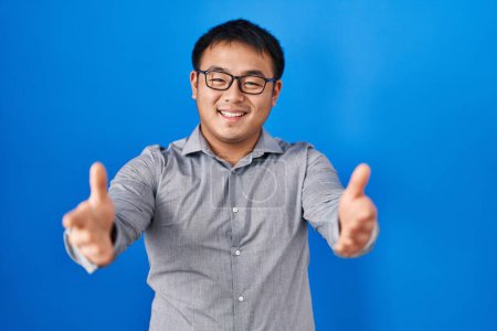 Téléchargez les photos : Jeune homme chinois debout sur fond bleu regardant la caméra souriant avec les bras ouverts pour étreindre. expression joyeuse embrassant le bonheur. - en image libre de droit