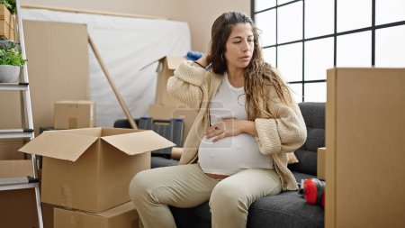 Téléchargez les photos : Jeune femme enceinte regardant autour de lui avec une expression sérieuse touchant le ventre à la nouvelle maison - en image libre de droit