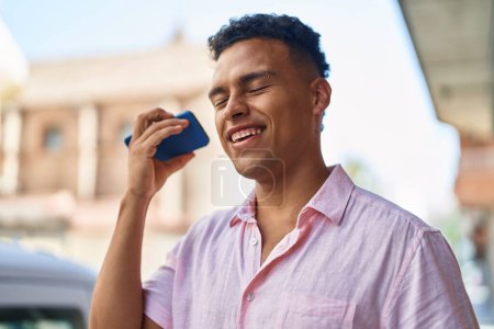 Téléchargez les photos : Jeune latin homme souriant confiant écoute message audio par le smartphone à la rue - en image libre de droit