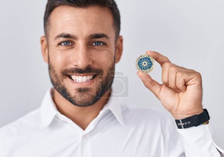Téléchargez les photos : Bel homme hispanique tenant pièce de monnaie crypto-monnaie cardano regardant positif et heureux debout et souriant avec un sourire confiant montrant dents - en image libre de droit