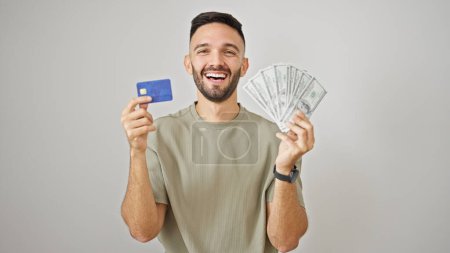 Foto de Joven hombre hispano sonriendo confiado sosteniendo tarjeta de crédito y dólares sobre fondo blanco aislado - Imagen libre de derechos