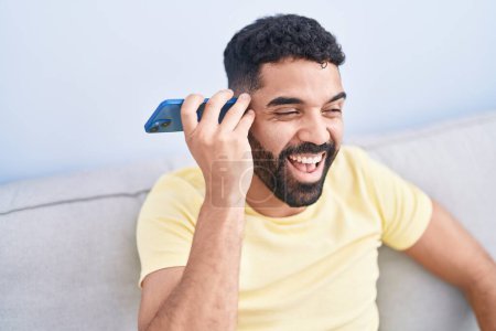 Téléchargez les photos : Young arab man listening voice message by smartphone sitting on sofa at home - en image libre de droit