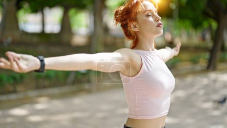 Photo for Young redhead woman wearing sportswear stretching arms breathing at park - Royalty Free Image