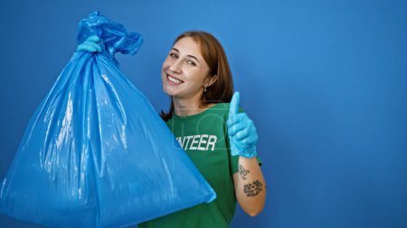 Téléchargez les photos : Jeune femme bénévole tenant sac poubelle faisant geste pouce vers le haut sur fond bleu isolé - en image libre de droit