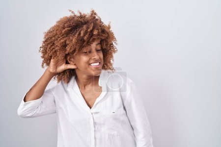 Photo for Young hispanic woman with curly hair standing over white background smiling with hand over ear listening an hearing to rumor or gossip. deafness concept. - Royalty Free Image