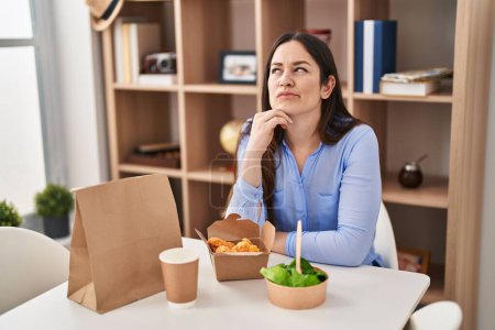 Téléchargez les photos : Jeune femme brune manger emporter de la nourriture à la maison visage sérieux penser à la question avec la main sur le menton, réfléchi sur l'idée déroutante - en image libre de droit