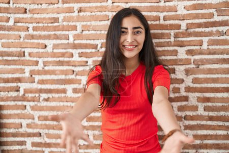 Téléchargez les photos : Jeune adolescente debout sur un mur de briques souriant joyeuse offrant des mains donnant assistance et acceptation. - en image libre de droit