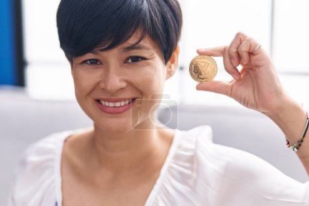 Foto de Joven mujer asiática con pelo corto sosteniendo moneda criptomoneda tron mirando positiva y feliz de pie y sonriendo con una sonrisa confiada mostrando los dientes - Imagen libre de derechos