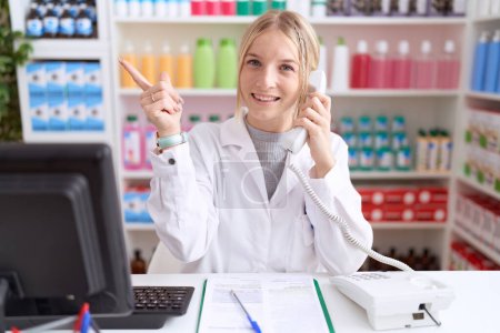 Téléchargez les photos : Jeune femme caucasienne travaillant à la pharmacie pharmacie parlant au téléphone avec un grand sourire sur le visage, pointant du doigt vers le côté en regardant la caméra. - en image libre de droit