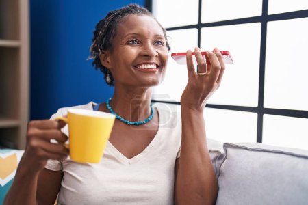 Téléchargez les photos : Moyen Age femme afro-américaine écoute un message vocal par smartphone boire du café à la maison - en image libre de droit