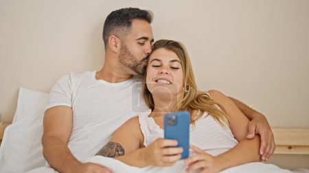 Téléchargez les photos : Homme et femme couple couché sur le lit à l'aide d'un smartphone se câlinant dans la chambre - en image libre de droit