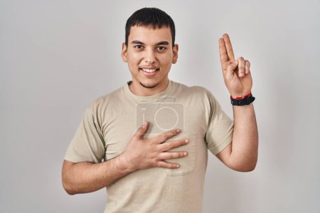 Téléchargez les photos : Jeune homme arabe portant un t-shirt décontracté souriant jurant avec la main sur la poitrine et les doigts vers le haut, faisant un serment de fidélité - en image libre de droit