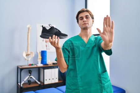 Téléchargez les photos : Jeune homme travaillant à la clinique de physiothérapie tenant la chaussure avec la main ouverte faisant stop signe avec expression sérieuse et confiante, geste de défense - en image libre de droit