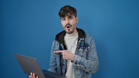 Foto de Joven hombre hispano señalando a la computadora portátil con la cara sorpresa sobre el fondo azul aislado - Imagen libre de derechos
