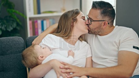 Téléchargez les photos : Famille de mère, père et bébé baisers assis sur le canapé à la maison - en image libre de droit