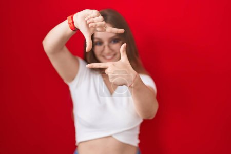 Téléchargez les photos : Jeune femme caucasienne debout sur fond rouge souriant faisant cadre avec les mains et les doigts avec le visage heureux. concept de créativité et photographie. - en image libre de droit