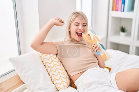 Photo for Young blonde woman singing song using brush as a microphone at bedroom - Royalty Free Image