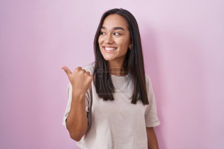 Foto de Young hispanic woman standing over pink background smiling with happy face looking and pointing to the side with thumb up. - Imagen libre de derechos
