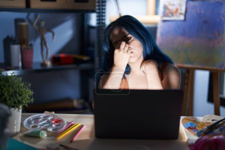 Foto de Joven chica moderna con el pelo azul sentado en el estudio de arte con el ordenador portátil por la noche cansada frotando la nariz y los ojos sintiendo fatiga y dolor de cabeza. concepto de estrés y frustración. - Imagen libre de derechos