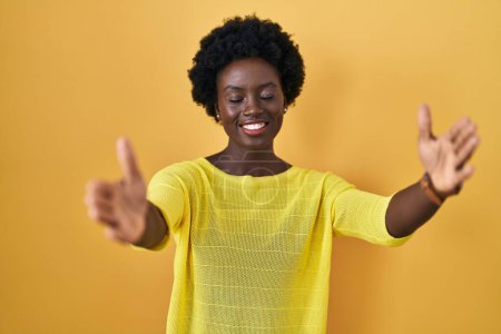 Foto de Mujer joven africana de pie sobre el estudio amarillo mirando a la cámara sonriendo con los brazos abiertos para abrazar. expresión alegre abrazando la felicidad. - Imagen libre de derechos
