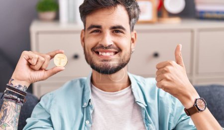 Photo for Young hispanic man with tattoos holding tether cryptocurrency coin smiling happy and positive, thumb up doing excellent and approval sign - Royalty Free Image