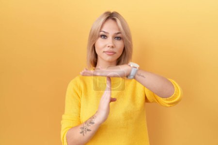 Foto de Mujer joven caucásica usando suéter amarillo haciendo el gesto del tiempo fuera con las manos, rostro frustrado y serio - Imagen libre de derechos