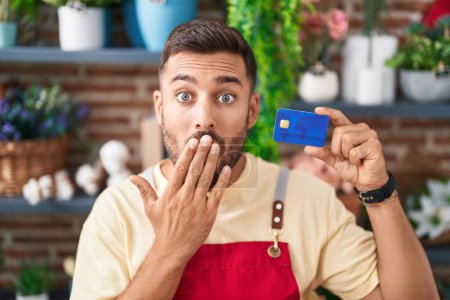Foto de Hombre hispano guapo que trabaja en floristería con tarjeta de crédito cubriendo la boca con la mano, sorprendido y temeroso de equivocarse. expresión sorprendida - Imagen libre de derechos