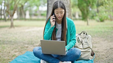Foto de Joven mujer china hablando en el teléfono inteligente utilizando el ordenador portátil en el parque - Imagen libre de derechos