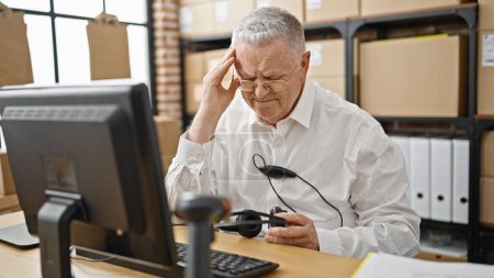 Foto de Hombre de pelo gris de mediana edad comercio electrónico trabajador de negocios cansado de sacar los auriculares que trabajan en la oficina - Imagen libre de derechos