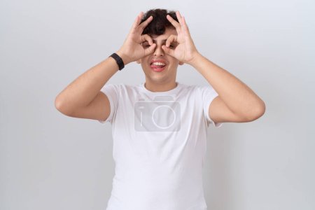 Foto de Joven hombre no binario con camiseta blanca casual haciendo buen gesto como binoculares sacando la lengua, ojos mirando a través de los dedos. expresión loca. - Imagen libre de derechos