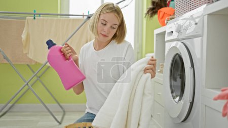 Photo for Young blonde woman holding clean towel and detergent bottle at laundry room - Royalty Free Image