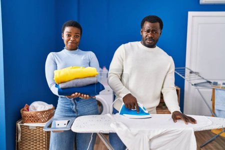 Foto de Joven pareja afroamericana planchando ropa en la lavandería expresión despistada y confusa. concepto de duda. - Imagen libre de derechos