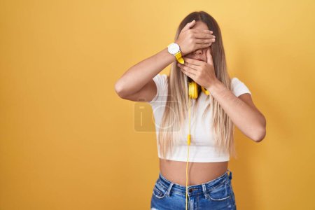 Foto de Mujer rubia joven de pie sobre fondo amarillo con auriculares que cubren los ojos y la boca con las manos, sorprendida y conmocionada. ocultar emociones - Imagen libre de derechos