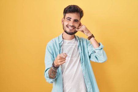 Photo for Young hispanic man with tattoos standing over yellow background smiling doing talking on the telephone gesture and pointing to you. call me. - Royalty Free Image