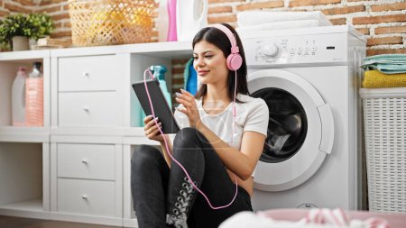 Foto de Mujer joven caucásica usando touchpad y auriculares esperando a la lavadora en la sala de lavandería - Imagen libre de derechos