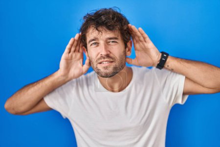 Photo for Hispanic young man standing over blue background trying to hear both hands on ear gesture, curious for gossip. hearing problem, deaf - Royalty Free Image