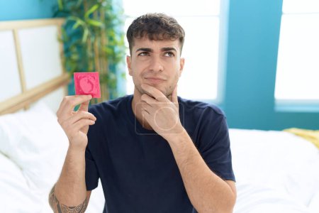 Téléchargez les photos : Jeune homme hispanique assis sur le lit tenant préservatif visage sérieux en pensant à la question avec la main sur le menton, réfléchi sur l'idée déroutante - en image libre de droit