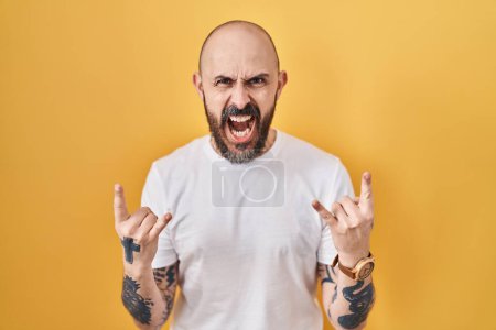Photo for Young hispanic man with tattoos standing over yellow background shouting with crazy expression doing rock symbol with hands up. music star. heavy music concept. - Royalty Free Image