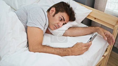 Foto de Hombre hispano joven usando teléfono inteligente acostado en la cama en el dormitorio - Imagen libre de derechos